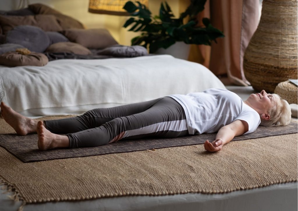 Kobieta w stroju sportowym leży na podłodze i wykonuje relaksujace ćwiczenia oddechowe.